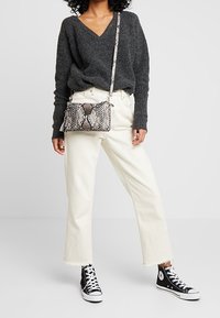 Borsa a tracolla in stampa serpente con superficie testurizzata, dotata di chiusura a zip e tracolla rimovibile. Indossata con maglione color carbone e pantaloni crema.