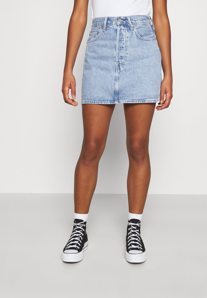 Femme portant une mini-jupe en denim bleu clair, des chaussettes blanches et des baskets montantes noires sur un fond blanc uni.