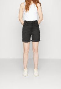 Shorts de mezclilla negros con puños enrollados, cintura de tiro medio, dos bolsillos delanteros y herrajes de cobre. Se llevan con una camiseta sin mangas blanca y zapatillas blancas.