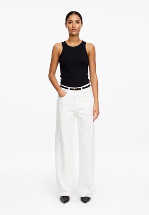 Kobieta stojąca w czarnym topie bez rękawów, białych, szerokich spodniach z wysokim stanem przewiązanych czarnym paskiem oraz w czarnych, spiczastych butach.