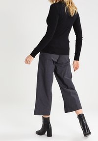 Femme portant un haut noir ajusté à manches longues, un pantalon court large gris et des bottines à talons noires, se tenant de dos.