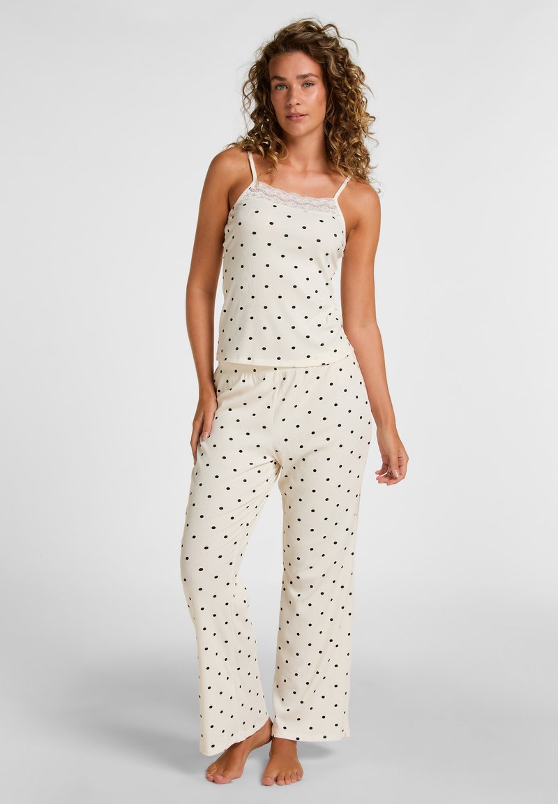 Femme aux cheveux bouclés portant un caraco crème et un pantalon à pois noirs, debout pieds nus devant un fond uni.