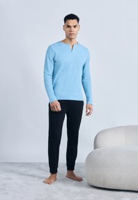 Άνδρας που στέκεται ξυπόλητος σε χαλί, φορώντας ανοιχτό μπλε μακρυμάνικη μπλούζα και μαύρο παντελόνι δίπλα σε καμπύλη λευκή υφασμάτινη καρέκλα.