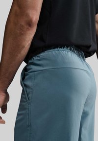 Herrebukser i lyseblåt stof med en glat tekstur, der har elastisk talje og sidelommer, set fra bagsiden.