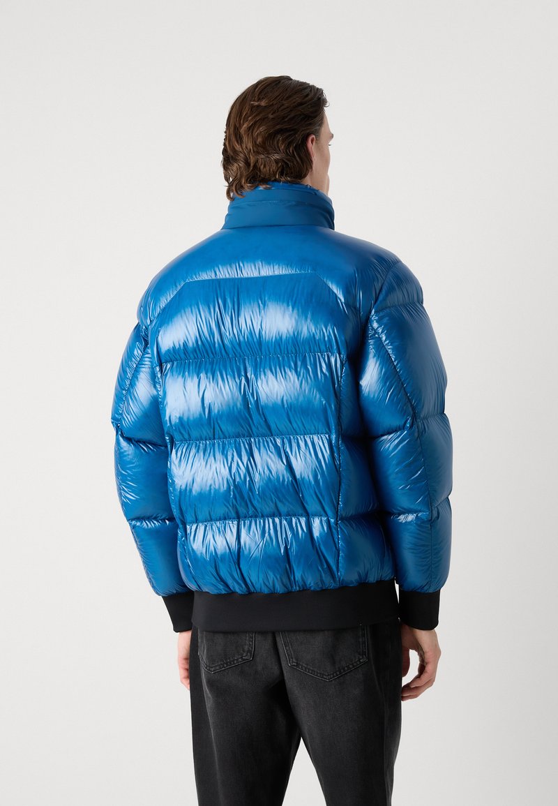 Veste rembourrée bleue avec une finition brillante, dotée d'un col montant, d'un design matelassé et de poignets élastiques. Associée à un jean noir pour créer du contraste.