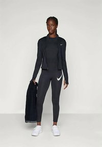 Tenue de sport noire comprenant une veste zippée, un haut ajusté et des leggings avec un swoosh blanc de Nike. Les baskets sont blanches ; le tissu semble lisse.