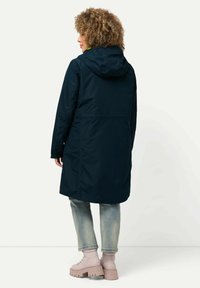 Giacca impermeabile di un blu navy scuro con cappuccio, vita cinchata e maniche lunghe, abbinata a jeans azzurri chiari e stivali rosa chunky.