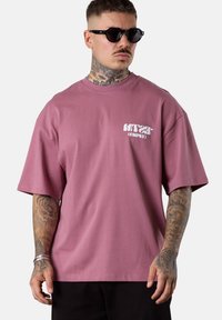 Mann mit schwarzen Sonnenbrillen und einem übergroßen mauvefarbenen T-Shirt mit weißem Text auf der Brust, der voll tätowierte Unterarme und einen tätowierten Hals zeigt.