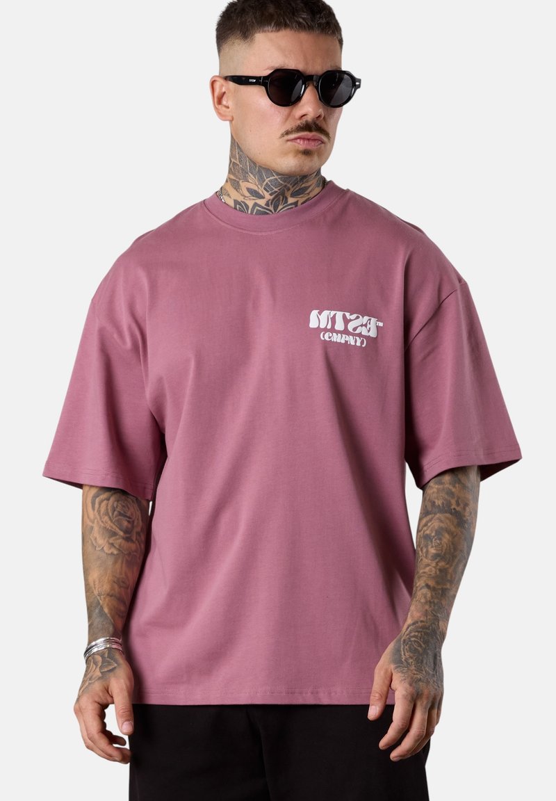 Mann mit schwarzen Sonnenbrillen und einem übergroßen mauvefarbenen T-Shirt mit weißem Text auf der Brust, der voll tätowierte Unterarme und einen tätowierten Hals zeigt.