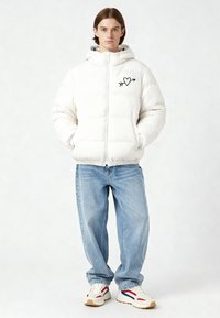 Giacca imbottita bianca con chiusura a zip, tasche anteriori e ricamo nero a forma di cuore e freccia. Abbinata a jeans azzurri e sneaker bianche.