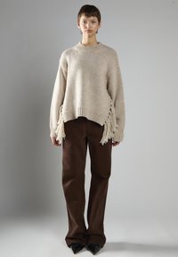 Osoba w beżowym swetrze z frędzlami, brązowych szerokich spodniach i czarnych szpiczastych butach, stojąca na tle plain szarego tła.