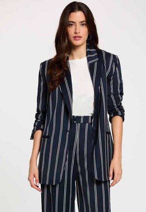 Mujer con cabello largo y oscuro, que lleva un traje azul marino con rayas verticales blancas y una blusa blanca, de pie frente a un fondo liso y claro.