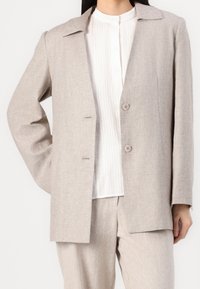 Hellbeige Blazer mit Reverskragen, Zwei-Knopf-Verschluss und subtiler Textur. Kombiniert mit einem weißen, gefalteten Hemd mit Knopfleiste darunter.