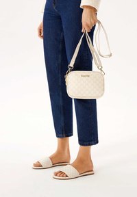 Borsa a tracolla bianca con motivo e hardware dorato, abbinata a sandali flat beige, indossata con jeans blu a vita corta.