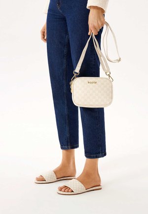 Borsa a tracolla bianca con motivo e hardware dorato, abbinata a sandali flat beige, indossata con jeans blu a vita corta.