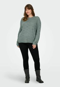 Ljusgrön stickad tröja med ett strukturerat mönster, rund halsringning, långa ärmar, kombinerad med svarta slim-fit-byxor och svarta ankelboots.