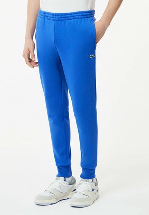 Personne portant un pantalon de jogging bleu vif avec une taille élastique et des baskets blanches avec des accents bleus, debout les mains près des poches.