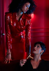 Zwei Models in einem roten, glänzenden Latex-Outfit und einem schwarzen, strukturierten Kleid, mit auffälligem Schmuck, in einem pink beleuchteten Innenraum.