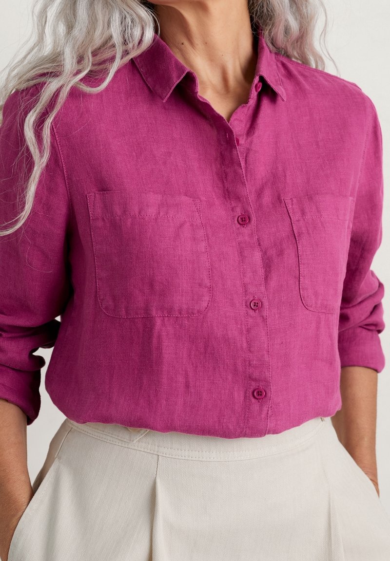 Kvinna som bär en magenta knapp-up skjorta med upprullade ärmar och två bröstfickor, instoppad i högmidjade beige byxor med händerna i fickorna.