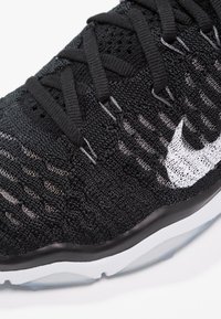 Scarpa atletica nera con tomaia in maglia, dotata di un logo bianco, motivo testurizzato e una suola nera con un accento bianco.