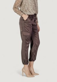 Pantaloni jogger in satin di colore marrone scuro con polsini elasticizzati e tasche laterali, abbinati a una blusa a fantasia e tacchi color carne.