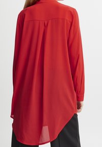 Camicia rossa a maniche lunghe con orlo curvo, realizzata in tessuto leggero, con piega sul retro e polsini con bottoni.