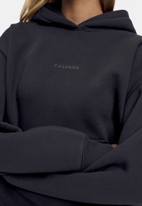 Mørk grå hettegenser laget av mykt stoff, med logo på forsiden og ribbestrikkede mansjetter. Den har en avslappet passform og foret hette.