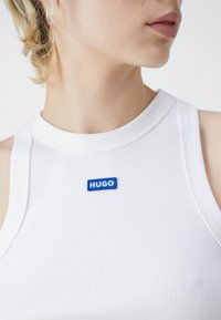 Vit ribbad linne med rund halsringning, med en blå broderad "HUGO" etikett på bröstet. Strukturerat tyg och figursydd design.