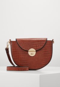 Borsa a tracolla marrone a mezzaluna con texture a rilievo coccodrillo, chiusura a girotondo dorata e tracolla regolabile su piedistallo bianco.