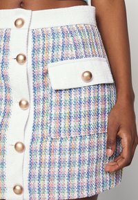 Jupe en tweed colorée avec une ceinture blanche et des boutons, présentant une poche avant avec un grand accent métallique. Détail du tissu texturé visible.