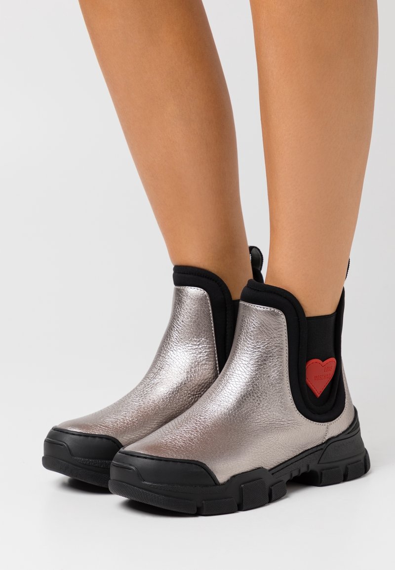 Bottines métalliques argentées avec semelles et garnitures noires, avec des patchs rouges en forme de cœur sur les côtés, portées sur des jambes nues sur fond blanc.