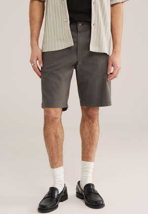 Mężczyzna ubrany w ciemnoszare krótkie spodenki za kolano, białe skarpetki, czarne skórzane mokasyny, czarną koszulę oraz jasnobeżową teksturowaną koszulę z krótkim rękawem zapinaną na guziki.