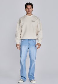 Beige katoenen sweatshirt met een ronde hals en een subtiel logo op de borst, gecombineerd met lichtblauwe denim flared jeans en witte sneakers.
