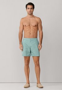 Hombre sin camisa de pie, vistiendo pantalones cortos de baño azul claro y zapatos sin cordones beige contra un fondo gris liso.
