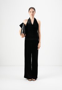 Vrouw die staat met een zwarte haltertop en wijde zwarte broek, met een grote zwarte clutch in de hand, tegen een witte achtergrond.