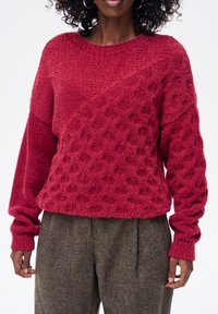 Czerwony dziergany sweter z teksturowanymi wzorami, okrągłym dekoltem i długimi rękawami. W zestawie z brązowymi teksturowanymi spodniami.