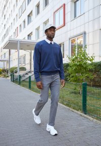 Marine sweatshirt met witte kraag, grijze jeans en witte sportschoenen. Wandelt op een stoep met een modern gebouw op de achtergrond.