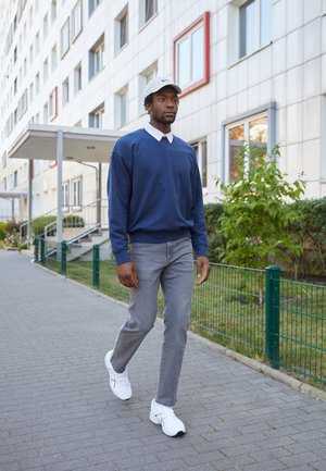 Sudadera navy con cuello blanco, jeans grises y zapatillas deportivas blancas. Caminando por una acera con un edificio moderno de fondo.