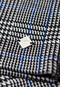 Tissu pied-de-poule noir et blanc avec des rayures bleues, doté d'une étiquette carrée en aluminium argenté portant l'inscription "Super Dry", fixée par des coutures noires.