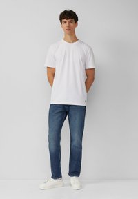 T-shirt bianco in cotone con scollatura rotonda, abbinato a jeans denim blu. Il modello indossa sneaker bianche e sta in piedi rivolto in avanti con le mani in tasca.