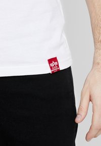 Hvit bomull t-skjorte med en rød etikett som har "Alpha Industries"-logoen, og svarte bukser som er delvis synlige nederst.