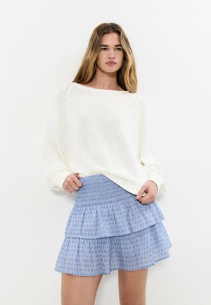 Jeune femme aux longs cheveux portant un pull en maille blanc et une mini-jupe bleue à volants avec un motif subtil sur un fond uni.