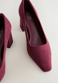 Scarpe con tacco alto in suede borgogna, con punta squadrata e tacco a blocco, caratterizzate da una texture liscia e un design minimalista.