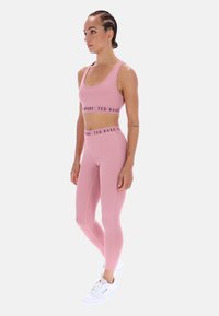 Világos rózsaszín sportmelltartó és hozzáillő, magas derekú leggings, sima anyagból, logó díszítéssel. Fehér sportcipővel párosítva.