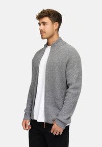 Cárdigan de punto gris con cremallera y cuello alzado, textura acanalada y mangas largas, llevado sobre una camiseta blanca y pantalones negros.