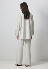 Chemise gris clair à manches retroussées, coupe ample et empiècement au dos ; associée à un pantalon large assorti, avec une texture minimale et des sandales plates.