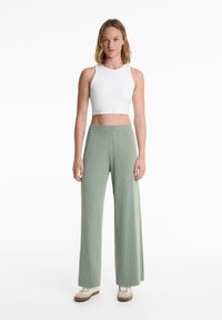 En stående kvinna bär en vit crop top och gröna byxor med vida ben, i kombination med beige sneakers, mot en enfärgad vit bakgrund.