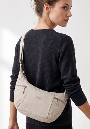 Vrouw met een donkere trui en een beige crossbodytas met meerdere ritsvakken en de letters "GMV" aan de voorkant.