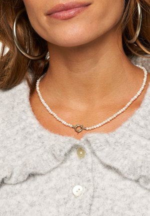 Pearl necklace featuring small white pearls with a metallic toggle clasp, worn with a gray, textured cardigan and oversized silver hoop earrings.