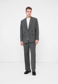 Sacou gri cu dungi fine, cu două rânduri de nasturi, asortat cu pantaloni din aceeași țesătură. Ținuta include un tricou alb și pantofi eleganți negri.